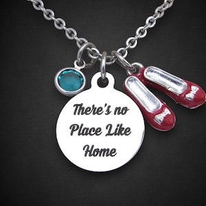 May include: Silver-toned necklace with a round pendant inscribed with "There's no Place Like Home." The necklace features a teal-colored charm and two small red ruby slipper charms. The chain is made of small, linked circles.