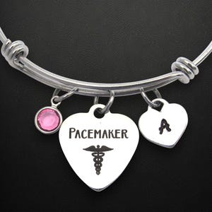 May include: A silver-toned bangle bracelet with three charms. One charm is a pink gemstone, another is a heart-shaped charm with the word "Pacemaker" and a medical symbol, and the third is a heart with the letter "A".