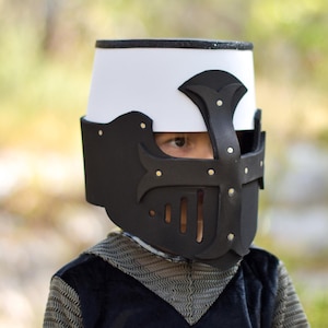 May include: A child's knight helmet, black and white with a cross design and gold rivets. The helmet has a white top and a black face guard with eye slits. The child is also wearing a chainmail shirt.