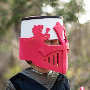 May include: A red and white knight's helmet featuring a lion emblem. The helmet has a white top with black trim and a red face guard with a cross design. The person is wearing a black shirt with chainmail.