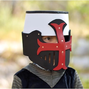 May include: A child wearing a toy knight's helmet. The helmet is white with a black visor and a red cross design. The helmet has gold-coloured rivets. The child is wearing a chainmail shirt.