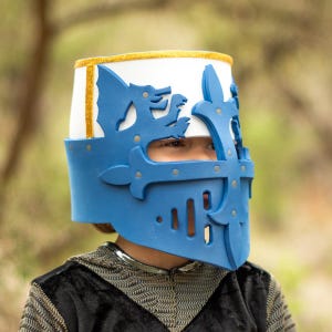 May include: A blue and white knight's helmet with gold trim and decorative cutouts. The helmet has a visor with eye slits and is worn over a black and silver garment. The helmet is designed for costume or play.