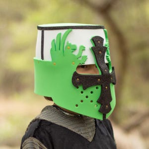 May include: A green and white toy helmet with a black cross and decorative accents. The helmet features a green dragon design and is worn by a person. The helmet is a costume piece.