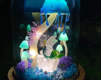 May include: A glass dome with a wooden base containing a miniature fairy garden. The garden features a blue and white mushroom with a glowing light, a fairy figurine, and other miniature plants and flowers.