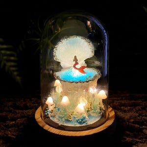 May include: A decorative glass dome with a miniature mermaid scene. The scene includes a mermaid figure, a seashell, and glowing mushrooms. The base is made of wood. The mermaid has a red tail and is set against a blue backdrop.