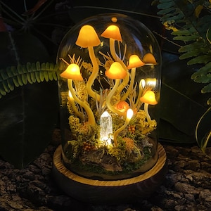 Puede incluir: Una c&uacute;pula de vidrio con una base de madera que contiene una escena de bosque en miniatura con hongos amarillos brillantes y un cristal transparente.