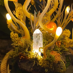 Puede incluir: Un cristal brillante se encuentra en un lecho de musgo y flores amarillas, rodeado de ramas de vidrio amarillo brillantes.
