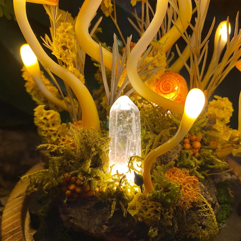 Puede incluir: Un cristal brillante se encuentra en un lecho de musgo y flores amarillas, rodeado de ramas de vidrio amarillo brillantes.