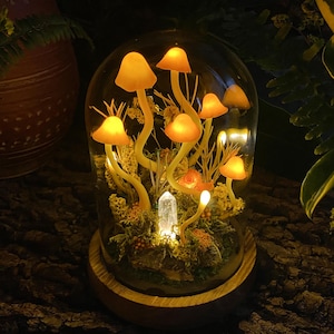 Puede incluir: Una c&uacute;pula de vidrio con una base de madera que contiene una escena de bosque en miniatura con hongos amarillos brillantes y un cristal transparente.