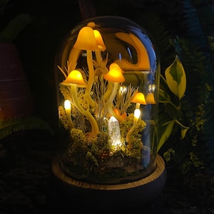 Puede incluir: Un terrario de c&uacute;pula de vidrio con una escena de bosque en miniatura que presenta hongos amarillos brillantes y un cristal transparente. La base del terrario est&aacute; hecha de madera.