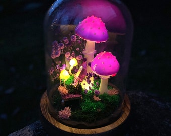 May include: A decorative glass dome featuring illuminated pink mushrooms with white stems, surrounded by purple flowers and moss. The base is made of wood. The scene is lit with small yellow lights, creating a whimsical, fairy-tale aesthetic.