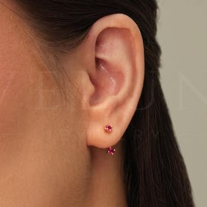 Pode incluir: Um par de brincos de ouro com pequenas pedras preciosas vermelhas. Os brincos são usados no lóbulo da orelha de uma pessoa com cabelo castanho.