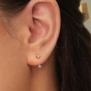 May include: A close-up of a person's ear with two small, gold stud earrings with tiny, clear gemstones.
