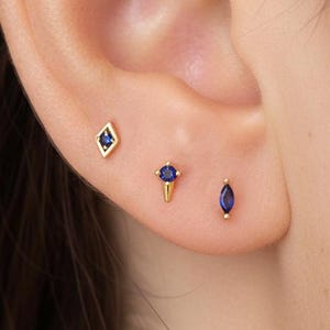 Peut inclure: Trois boucles d'oreilles dorées avec des pierres bleues sont présentées sur une oreille. Les boucles d'oreilles comprennent une forme de losange, une petite forme de cône et une forme ovale. Les boucles d'oreilles sont petites et délicates.