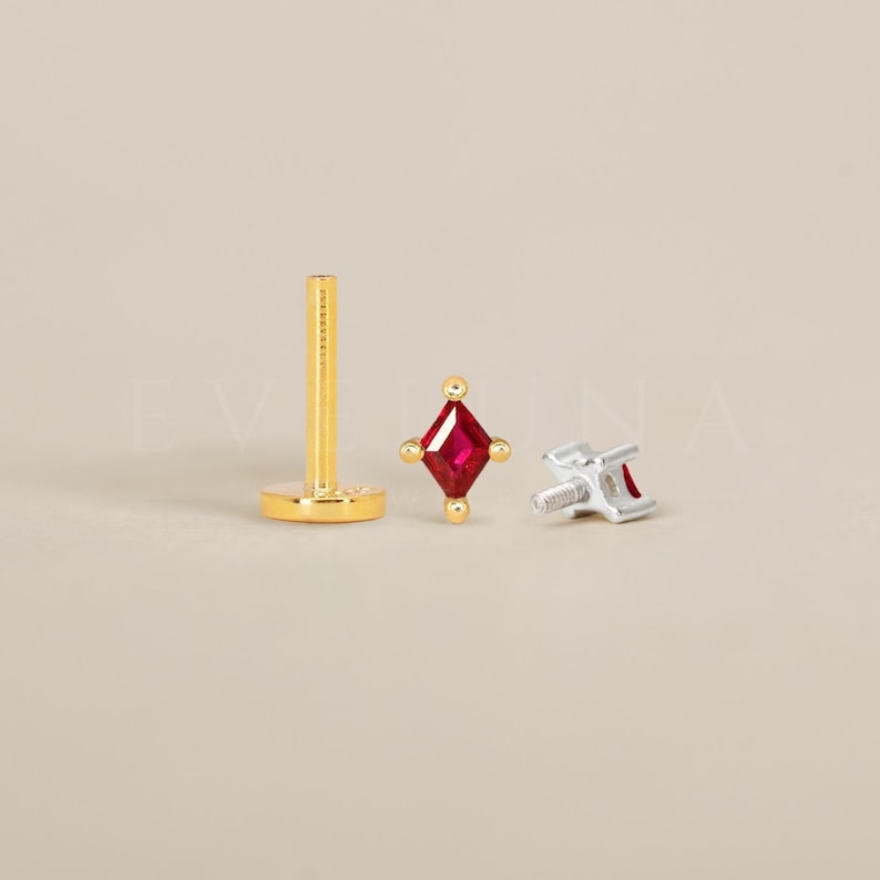 Może przedstawiać: Złoty kolczyk do nosa z małym, kwadratowym, czerwonym kamieniem.