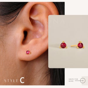 May include: A pair of gold stud earrings with small red gemstones. The earrings are shown on a model's ear. The earrings are simple and elegant.