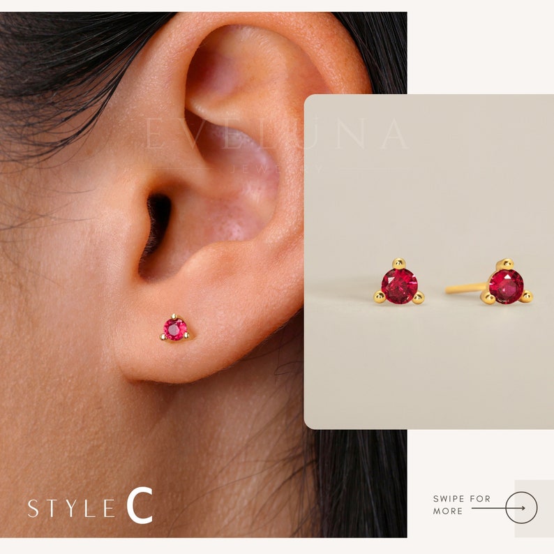 May include: A pair of gold stud earrings with small red gemstones. The earrings are shown on a model's ear. The earrings are simple and elegant.