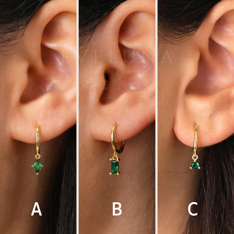 May include: Gold hoop earrings with emerald green gemstones in three different shapes: a square, a rectangle, and a teardrop. The earrings are shown on an ear, with the brand name "EVELUNA JEWELRY" visible in the background.