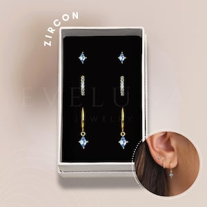 May include: A set of three pairs of earrings in a white box. The earrings are gold-toned and feature blue cubic zirconia stones. The box has the text "ELUA JEWELRY" printed on it.