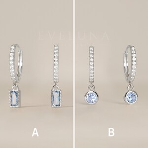 May include: Two pairs of silver hoop earrings with a dangling charm. The first pair has a rectangular blue gemstone charm. The second pair has a round blue gemstone charm.