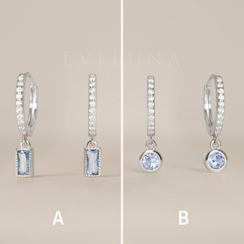 May include: Two pairs of silver hoop earrings with a dangling charm. The first pair has a rectangular blue gemstone charm. The second pair has a round blue gemstone charm.