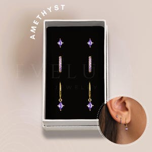 Puede incluir: Un conjunto de pendientes de amatista en una caja blanca. El conjunto incluye un par de pequeños pendientes de botón cuadrados, un par de pendientes de aro con pequeñas piedras de amatista y un par de pendientes colgantes con una piedra de amatista más grande.