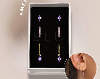 May include: A set of amethyst earrings in a white box. The set includes a pair of small square stud earrings, a pair of hoop earrings with small amethyst stones, and a pair of drop earrings with a larger amethyst stone.