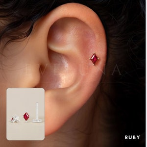 Może przedstawiać: Zbliżenie na kolczyk z rubinem. Kolczyk to mały, kwadratowy kamień szlachetny osadzony w złotej oprawie. Kolczyk jest noszony w g&oacute;rnej części chrząstki ucha.