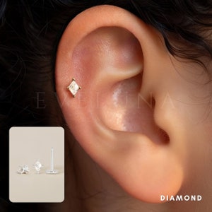 Może przedstawiać: Zbliżenie ucha z małym, kwadratowym kolczykiem z diamentem. Kolczyk jest osadzony w złotej oprawie i widoczny w g&oacute;rnym płatku ucha. Kolczyk ma prosty i elegancki design.