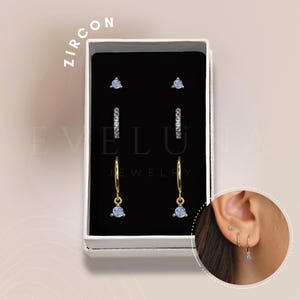 May include: A set of three gold hoop earrings with small blue zircon stones. The earrings are packaged in a white box with the word "ZIRCON" printed on the top. The box also has the words "EVELU JEWELRY" printed on the front.