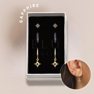 Może przedstawiać: Zestaw złotych kolczyków z niebieskimi szafirowymi akcentami. Kolczyki występują w trzech stylach: mała gwiazdka, obręcz z niebieskimi szafirami i obręcz z wiszącą gwiazdką. Kolczyki są zapakowane w białe pudełko z nadrukiem "Sapphire" i "Elu Jewelry".