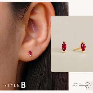 May include: A pair of gold earrings with small red marquise-shaped gemstones. The earrings are shown on a model's ear.