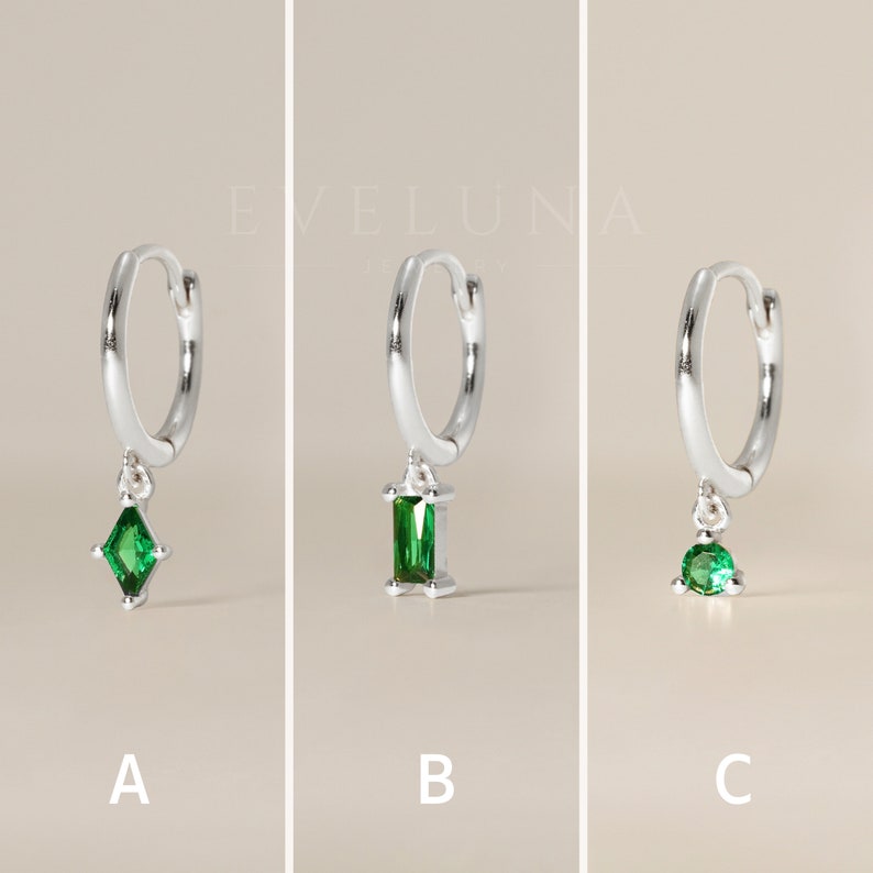 May include: Three silver hoop earrings with different green gemstone pendants. The earrings are labeled A, B, and C. Each earring has a different gemstone shape: a diamond, a rectangle, and a round shape. The brand name "EVELUNA JEWELRY" is in the background.
