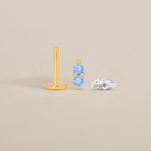Könnte beinhalten: Ein goldfarbener Labret-Stecker mit flachem Boden und Stift, neben einem goldenen Stecker mit zwei hellblauen Edelsteinen und einem silberfarbenen Rücken. Der Schmuck ist vor einem neutralen Hintergrund platziert.