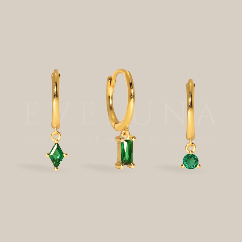 May include: Three gold-colored hoop earrings with dangling green gemstones. One earring features a diamond-shaped gem, another a rectangular gem, and the third a round gem. The earrings are displayed against a neutral background.
