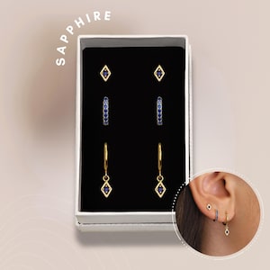 May include: A set of three pairs of gold earrings with blue sapphire stones. The earrings are in a white box with the word "SAPPHIRE" printed on the top. The box is open and the earrings are visible. The earrings are a pair of small studs, a pair of small hoops, and a pair of dangle earrings.