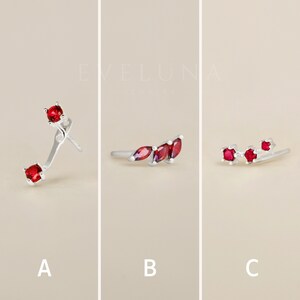 Op de afbeelding: Drie verschillende stijlen oorbellen met rode edelstenen. De eerste oorbel heeft een enkele rode edelsteen op een gebogen zilveren pen. De tweede oorbel heeft drie rode edelstenen in een gebogen zilveren setting. De derde oorbel heeft drie rode edelstenen in een rechte zilveren setting.