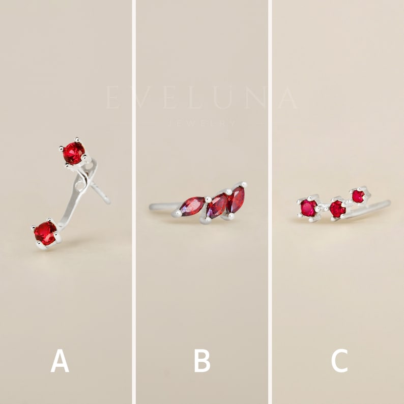 Op de afbeelding: Drie verschillende stijlen oorbellen met rode edelstenen. De eerste oorbel heeft een enkele rode edelsteen op een gebogen zilveren pen. De tweede oorbel heeft drie rode edelstenen in een gebogen zilveren setting. De derde oorbel heeft drie rode edelstenen in een rechte zilveren setting.