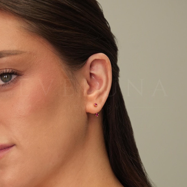 Op de afbeelding: Een paar gouden oorbelletjes met kleine rode edelstenen. De oorbelletjes worden gedragen in de oorlel van een persoon met bruin haar.