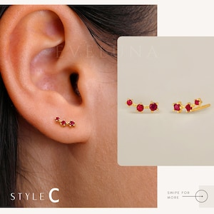 Op de afbeelding: Een paar gouden oorbellen met elk drie rode edelstenen. De oorbellen hebben de vorm van een gebogen lijn. De oorbellen worden getoond op een oormodel.