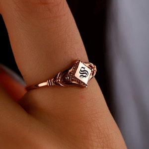 May include: A rose gold ring with a diamond-shaped setting featuring the letter "S" in a black font.