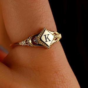 May include: A gold ring with a diamond-shaped setting featuring the letter "K" in the center. The ring has a delicate, ornate design.