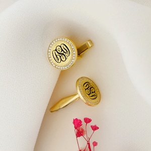 May include: A pair of gold-colored cufflinks with a monogram design. Each cufflink features a round face with a black monogram, surrounded by a ring of small, clear stones. The cufflinks are set against a white background.