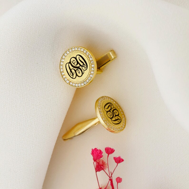 May include: A pair of gold-colored cufflinks with a monogram design. Each cufflink features a round face with a black monogram, surrounded by a ring of small, clear stones. The cufflinks are set against a white background.