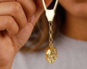 May include: A gold-toned metal keychain featuring a polished, teardrop-shaped ring attached to a short, delicate chain link. At the end of the chain hangs a small, detailed three-dimensional skull charm with hollow eye sockets and a defined jawline.