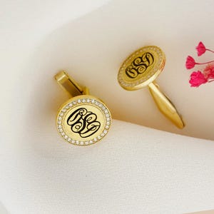 May include: Gold-toned cufflinks with a monogram design. Each cufflink features a circular face with a black monogram, surrounded by a ring of small, clear stones. The cufflinks are set against a white fabric background.