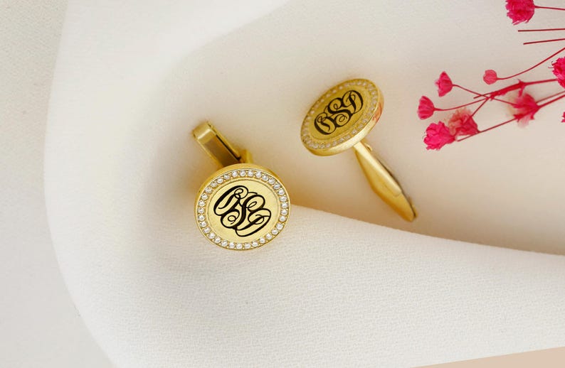 May include: Gold-toned cufflinks with a monogram design. Each cufflink features a circular face with a black monogram, surrounded by a ring of small, clear stones. The cufflinks are set against a white fabric background.