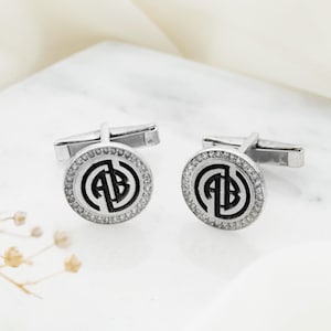 May include: A pair of silver cufflinks with a circular design. Each cufflink features a black monogram within a circle of clear crystals. The cufflinks are displayed on a white surface with dried flowers.