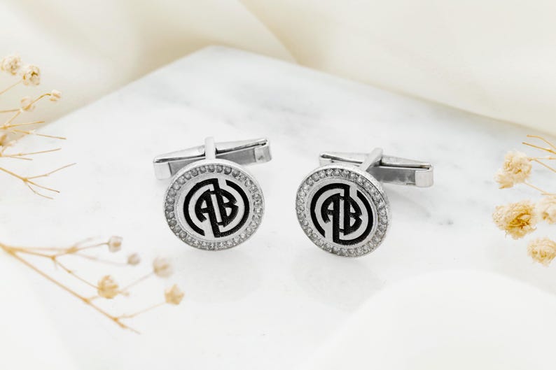 May include: A pair of silver cufflinks with a circular design. Each cufflink features a black monogram within a circle of clear crystals. The cufflinks are displayed on a white surface with dried flowers.