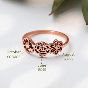 May include: A rose gold ring with a delicate floral design. The ring features three flowers, a rose, a poppy, and a cosmos. The flowers are arranged in a row and are connected by a thin band. The ring is a beautiful and unique piece of jewelry.